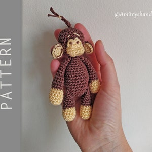 Könnte beinhalten: Ein gehäkeltes Affenspielzeug mit braunem Fell und gelben Händen und Füßen. Der Affe hat einen langen Schwanz und hält ein kleines, braunes Objekt in seiner Hand. Das Spielzeug wird in der Hand einer Person gehalten.