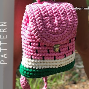 Puede incluir: Una mochila de sandía de crochet rosa, blanca y verde con una correa negra. La mochila tiene una solapa pequeña con un cierre plateado.