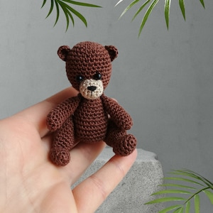May include: A small brown crocheted teddy bear with black eyes and a beige nose. The bear is sitting in a person's hand.