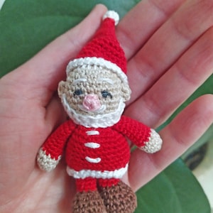 Op de afbeelding: Een klein gehaakt kerstman ornament met een rode muts, witte baard en bruine laarzen.