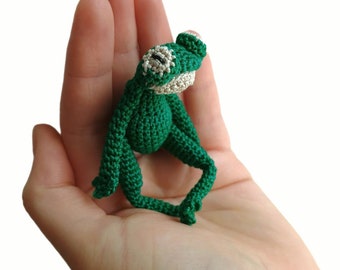 May include: A small, handmade green crochet frog with white accents on its eyes and mouth. The frog has long legs and is posed as if sitting. The frog is a whimsical, handcrafted item.