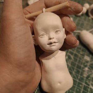 May include: A white, unfinished doll sculpture held by a hand. The doll has a detailed face with visible eyes, nose, and lips. The body is also sculpted, showing the torso and part of the arms. A wooden dowel is attached to the head.