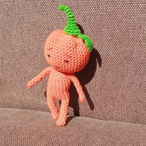 May include: A crocheted orange pumpkin-shaped character with a green stem and a sad expression. The character has long arms and legs and is standing on a brown textured surface.