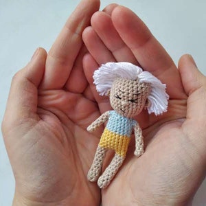 Puede incluir: Una pequeña muñeca de crochet con pelo blanco, una camisa azul y amarilla y una expresión de ojos cerrados.