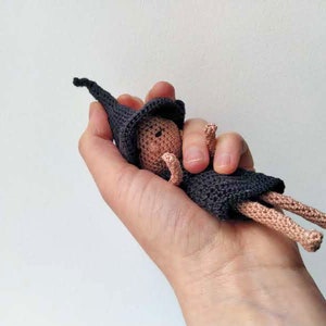 Può includere: Una piccola bambola all'uncinetto che indossa un cappello nero a punta e un vestito nero. La bambola ha un'incarnato marrone chiaro ed è sdraiata sulla schiena.