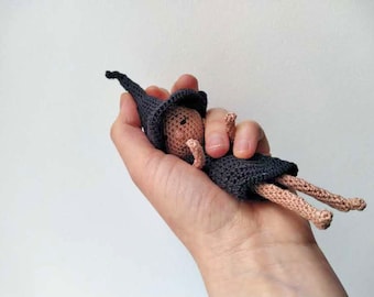 May include: A small crocheted doll wearing a black pointed hat and a black dress. The doll has a light brown complexion and is lying on its back.