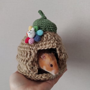 Puede incluir: Un pequeño hámster en una casa de ganchillo en forma de bellota con un diseño floral colorido. La casa está hecha de hilo marrón y verde.
