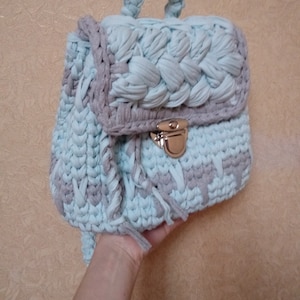 May include: A light blue and gray crocheted backpack with a silver clasp. The backpack has a braided handle and fringe details.
