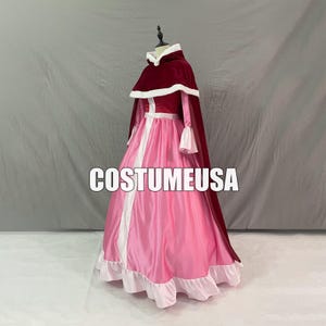 Könnte beinhalten: Ein rosa-weißes Kostümkleid mit burgunderfarbener Kapuze und Umhang. Das Kleid hat einen langen Rock mit weißem Besatz und ein Oberteil mit weißen Akzenten. Der Umhang ist mit weißem Kunstfell gefüttert. Der Text "COSTUMEUSA" steht auf der Vorderseite.