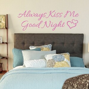 May include: A light blue bed with a brown patterned throw blanket. The headboard is dark brown with tufted buttons. The wall behind the bed has a pink vinyl decal that reads "Always Kiss Me Good Night" with two pink hearts.