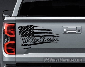 May include: A black vinyl decal applied to the silver tailgate of a pickup truck. The design features a distressed American flag graphic with horizontal brushstroke stripes and the text We the People in a stylized, traditional script font.