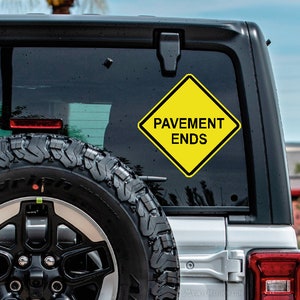 May include: A yellow diamond-shaped warning sign with black lettering that reads "Pavement Ends". The sign is attached to the back window of a silver SUV.