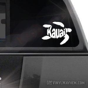May include: White vinyl decal of a sea turtle with the word "Kauai" spelled out within the turtle's body. The decal is on a black car window.