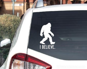 May include: A white vinyl decal applied to the rear window of a white vehicle. The decal features a solid white silhouette of the creature known as Bigfoot in a walking pose, positioned above the text "I BELIEVE." in a bold, sans-serif font.