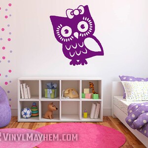 May include: Purple owl wall decal with a bow on its head. The owl is in a stylized design with large eyes and a patterned body. The decal is on a white wall in a child's bedroom.