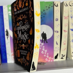 Op de afbeelding: Een hardcover boek met de titel "Combustible Magic" van Iris Beaglehole, onderdeel van de Myrtlewood Mysteries-serie. De omslag is zwart met gouden illustraties. De rand van het boek is crèmekleurig met gele illustraties. Andere boeken zijn zichtbaar op een plank.