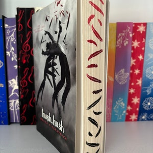 May include: A paperback book titled "Hush, Hush" by Becca Fitzpatrick. The cover features a black and white illustration of a person falling with feathers around them. The spine has a red and black feather design.