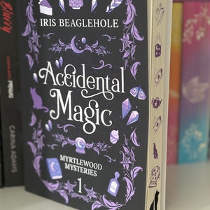 May include: A black book with a purple and white design featuring a teapot, a pie, and a crescent moon. The title is "Accidental Magic" and the author is Iris Beaglehole. The book is part of the Myrtlewood Mysteries series, book 1.