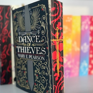 Könnte beinhalten: Ein Hardcover-Buch mit dem Titel "Dance of Thieves" von Mary E. Pearson. Das Buch hat einen dunkelblauen Einband mit goldenen und silbernen Akzenten sowie roten und goldenen floralen Kanten. Weitere Bücher sind im Hintergrund sichtbar.