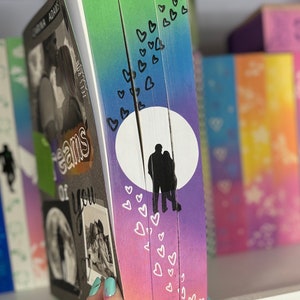 Könnte beinhalten: Ein Buch mit einem Regenbogen-Ombre-Umschlag und einer Silhouette eines Paares, das unter dem Mond steht. Der Umschlag ist mit weißen Herzen verziert.