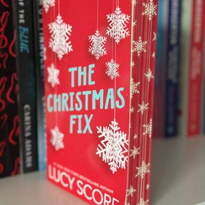 May include: A red paperback book with a white snowflake design on the cover. The title of the book is "The Christmas Fix" by Lucy Score.