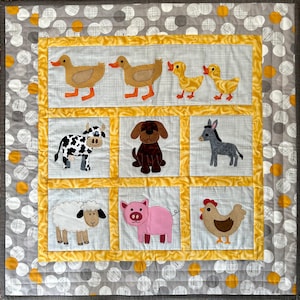 May include: A yellow and grey quilted fabric wall hanging with a white background. The fabric features a variety of embroidered animals, including a cow, a dog, a donkey, a pig, a chicken, a sheep, and two ducks.