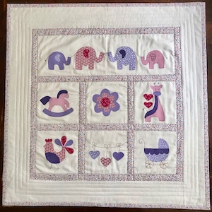 May include: A white baby quilt with a patchwork design featuring various animal and baby-themed appliques. The appliques are in shades of pink, purple, and white. The quilt is surrounded by a white border with a small, colourful polka dot pattern.