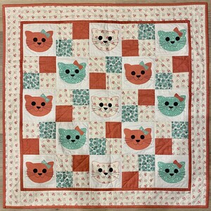 May include: A quilt with a patchwork design featuring various colored squares with a cat face design in each square. The cat faces are in various colors, including pink, green, and orange. The quilt is made with a variety of fabrics, including floral prints and solid colors.
