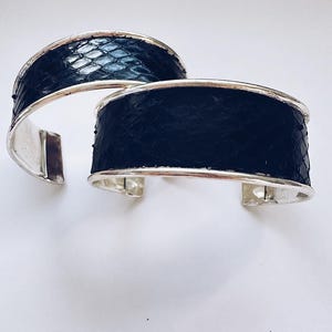 May include: Two silver bangle bracelets with black leather bands. The leather has a snakeskin pattern.