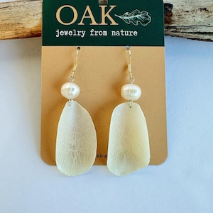 Puede incluir: Un par de pendientes de perlas blancas con un delicado diseño de hoja natural. Los pendientes están en una tarjeta que dice "OAK jewelry from nature".