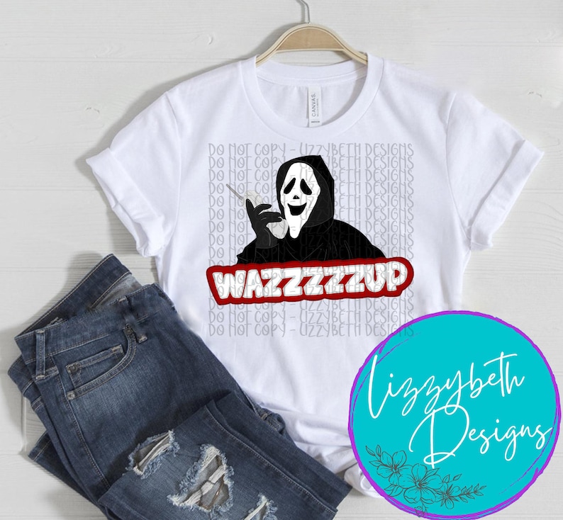 Op de afbeelding: Wit T-shirt met een zwart-witte illustratie van een gemaskerd figuur dat een telefoon vasthoudt en de tekst "WAZZZZZUP" in rood. De tekst "Lizzybeth Designs" staat in een cirkel met een blauwe achtergrond en witte bloemen.