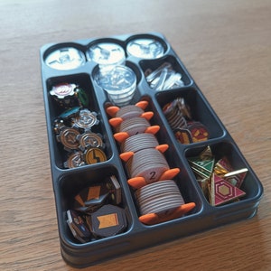 Puede incluir: Una bandeja organizadora de plástico negro llena de varias piezas de juego. La bandeja contiene piezas de plástico transparente, pilas de fichas de madera con detalles en naranja y componentes de juego de metal y cartón.