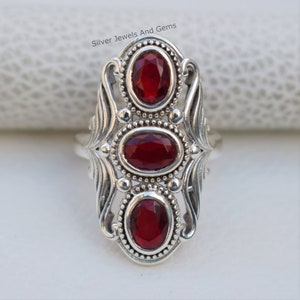 Puede incluir: Un anillo de plata con tres piedras preciosas rojas engastadas en un diseño ornamentado. El anillo tiene un diseño delicado e intrincado con un motivo floral.