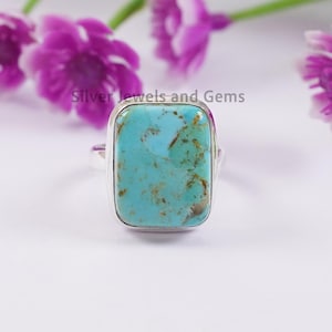May include: A silver ring with a large, rectangular turquoise stone. The turquoise has a mottled blue-green color with brown veining. The ring is set against a white background with purple flowers.