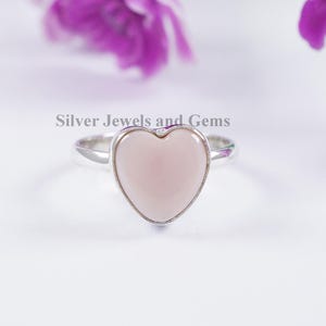 Peut inclure: Une bague en argent avec une pierre rose clair en forme de cœur, montée sur un anneau en métal poli. Le texte Silver Jewels and Gems est imprimé au-dessus de la bague. Le design est simple, mettant en valeur le cœur et l'anneau métallique.