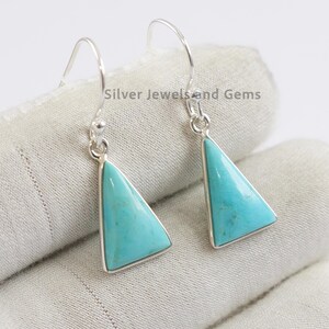 Puede incluir: Pendientes de plata con piedras triangulares turquesas. Los pendientes tienen un diseño de gancho y están colocados sobre un fondo claro. El texto "Silver Jewels and Gems" es visible.