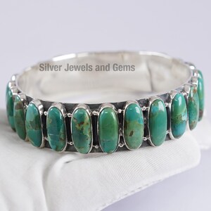 Könnte beinhalten: Ein silbernes Armband mit ovalen Türkissteinen. Die Steine haben eine leuchtende grün-blaue Farbe mit braunen Matrixmustern. Das Armband ist auf einem silbernen Band gefasst und wird auf einer weißen Oberfläche präsentiert. Der Text "Silver Jewels and Gems" ist sichtbar.