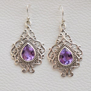 May include: A pair of silver drop earrings with a deep purple, pear-cut gemstone in each. The gemstones are set in ornate, filigree-style frames with a beaded edge and scrollwork. The earrings have a hook fastening.