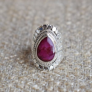 May include: A silver ring featuring a large, teardrop-shaped, pink ruby gemstone set in an intricate silver design. The ring is adorned with small, round silver beads.