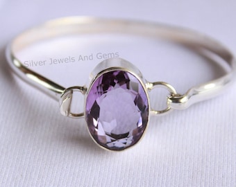 Könnte beinhalten: Ein silbernes Armreif-Armband mit einem großen ovalen Amethyst-Edelstein. Das Armband ist offen und hat ein schlichtes Design.
