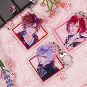 May include: Three colorful anime character keychains with red, pink, and blue accents. Each keychain features a different character with a unique design and is framed in a square shape.