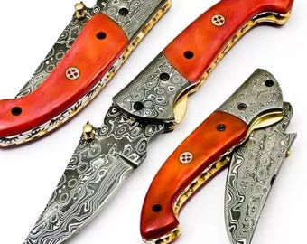 May include: Close-up of three folding knives. The knives have blades with a patterned design and orange handles. The handles are accented with gold-colored metal and small, decorative rivets. The blades are partially open, showcasing the intricate patterns.