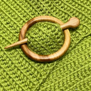 Op de afbeelding: Een lichtgroene gebreide stof met een ronde houten sjaalspeld. De speld bestaat uit een gladde, gepolijste houten ring met een bijpassende houten stok die door het midden gaat en het gestructureerde, limoengroene garenmateriaal op zijn plek houdt.