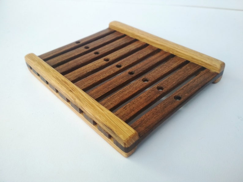 May include: A wooden soap dish with a slatted design. The dish is made of two types of wood, one light brown and one dark brown. The dish has several drainage holes.