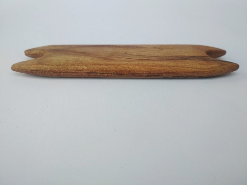 May include: A wooden tool with a rounded, slightly curved shape and two pointed ends. The tool is made of light brown wood with darker brown streaks.