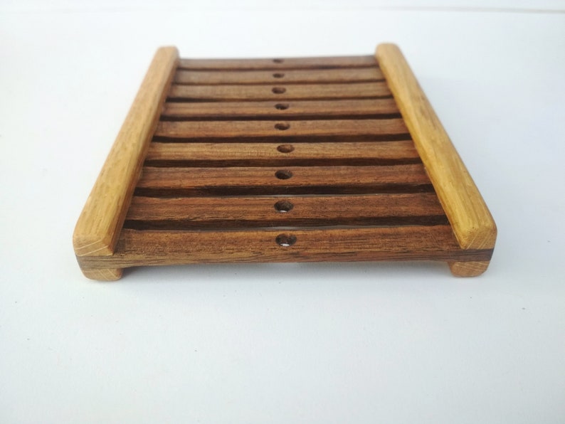 May include: A wooden soap dish with a slatted design. The dish is made of dark brown wood with lighter brown wood accents. The dish has a rectangular shape and is approximately 6 inches long and 4 inches wide.