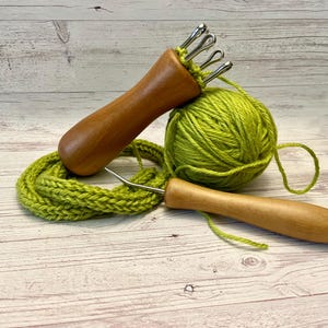 Puede incluir: Una herramienta de tejido de madera con múltiples agujas y una bola de hilo verde. El hilo está envuelto alrededor de la herramienta y un gancho de ganchillo de madera.
