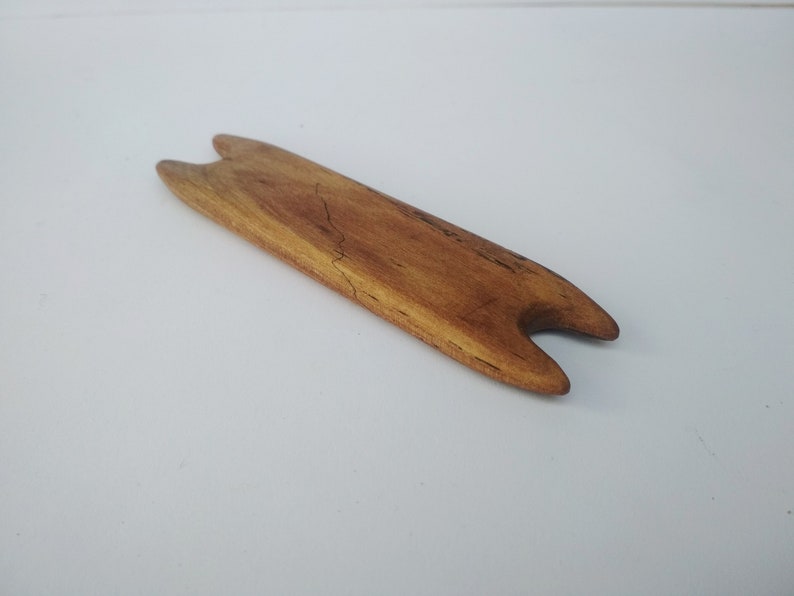 May include: A wooden knitting needle tool with two prongs at each end. The tool is light brown and has a natural wood grain.