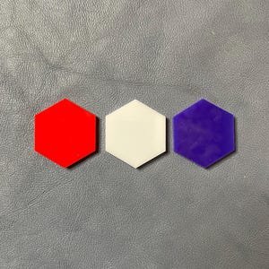 Puede incluir: Tres formas hexagonales rojas, blancas y moradas están dispuestas en fila. Las formas son de material sólido y están colocadas sobre una superficie gris. Vista superior.