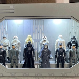 Può includere: Una vetrina rettangolare grigia con illuminazione interna, contenente una collezione di dieci action figure di Star Wars disposte su due file. Le figure includono vari stormtrooper, ufficiali, Darth Vader e l'Imperatore all'interno.